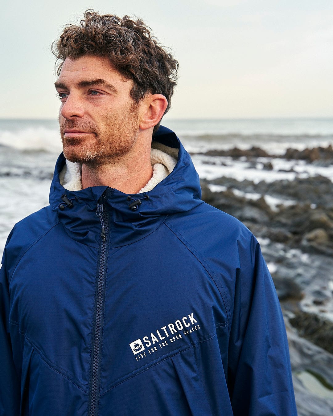 A person with short curly hair wearing a hooded blue Saltrock Changing Robe, adorned with the Saltrock logo, stands near a rocky shoreline with waves in the background.