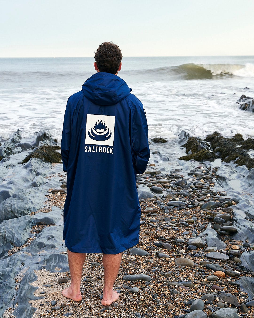 Person in a blue Saltrock Changing Robe - Blue adorned with the Saltrock logo stands barefoot on a rocky beach, facing the ocean waves.