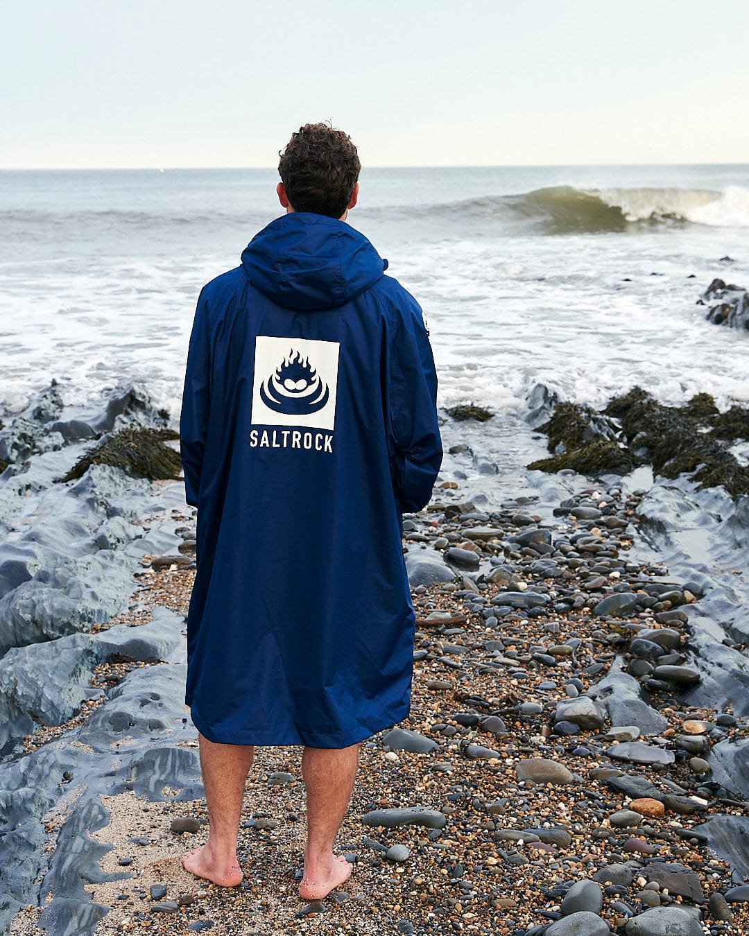 Man in a Saltrock Recycled Four Seasons Changing Robe - Blue standing on a pebble beach, looking at the ocean waves.