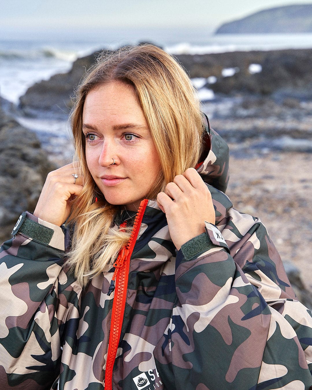 A woman in a Recycled Four Seasons Changing Robe - Brown Camo designed by Saltrock on a beach, adjusting the hood around her head.