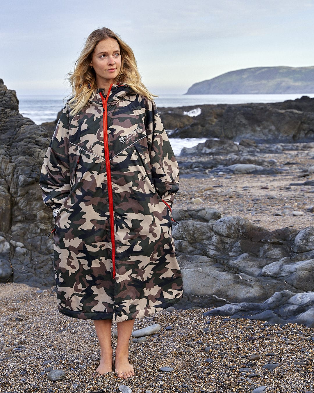 Woman in a Saltrock Recycled Four Seasons Changing Robe - Brown Camo standing on a rocky beach with the ocean in the background.