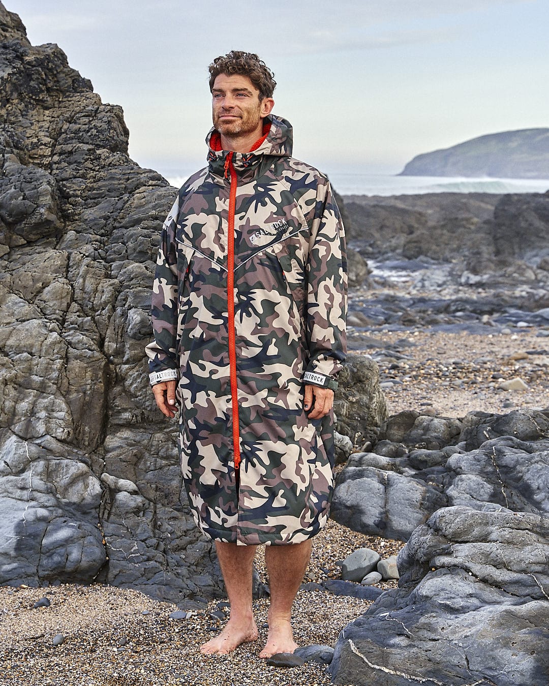 A man in a Saltrock camouflage raincoat, Four Seasons - Waterproof Changing Robe - Brown, standing on a rocky beach.