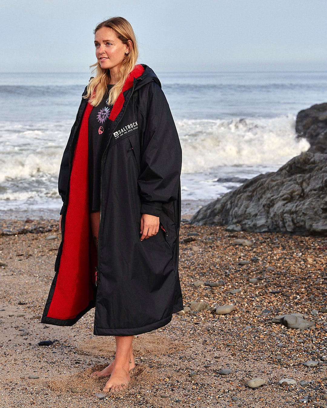 A person stands barefoot on a rocky beach wearing a long black and red coat with the words "Recycled Changing Robe - Black/Red" visible on the left side, made from recycled materials by Saltrock. The ocean and waves crash in the background.