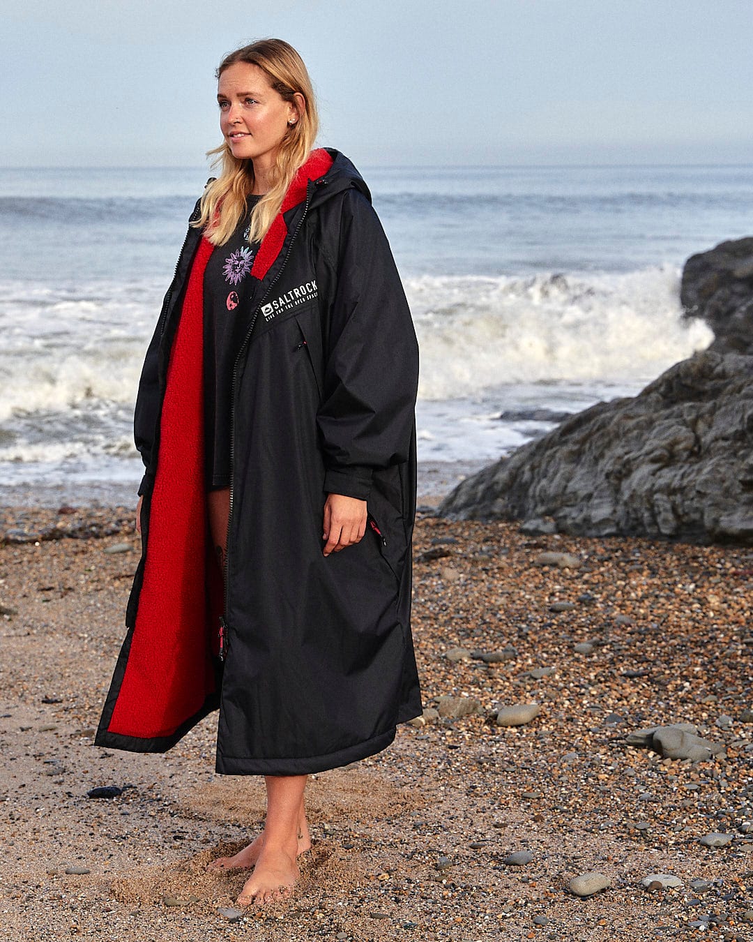 Woman in a Saltrock Recycled Four Seasons Changing Robe - Black/Red standing on a beach with waves and rocks in the background.