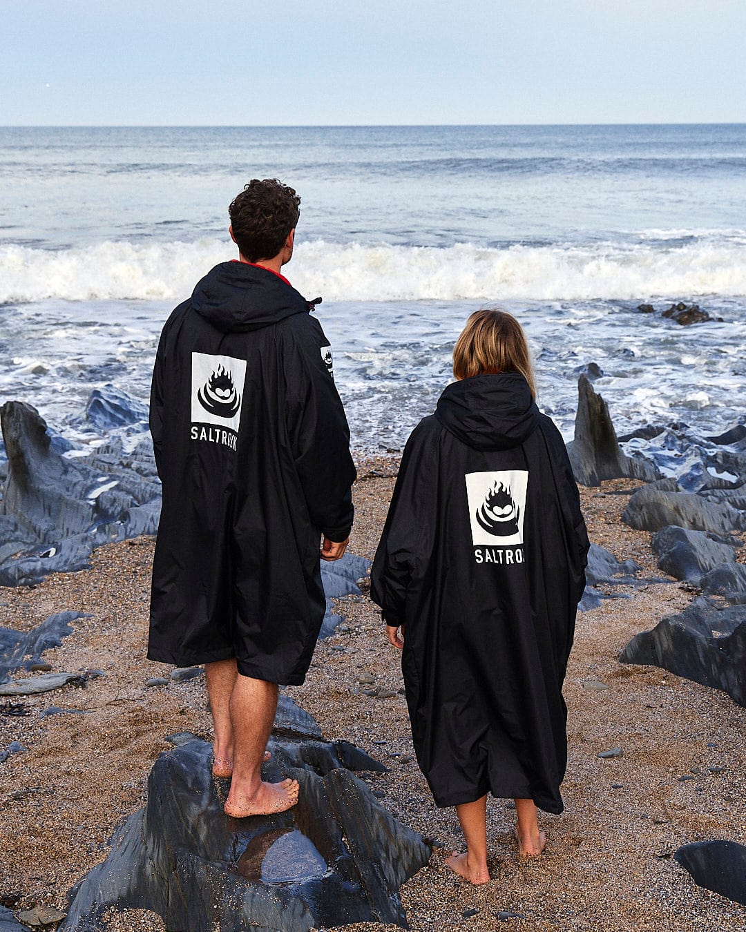 Two people standing on a rocky beach, facing the ocean in their black Recycled Changing Robes - Black/Red with the Saltrock logo on the back, crafted from recycled materials. The waves gently crash on the shore.