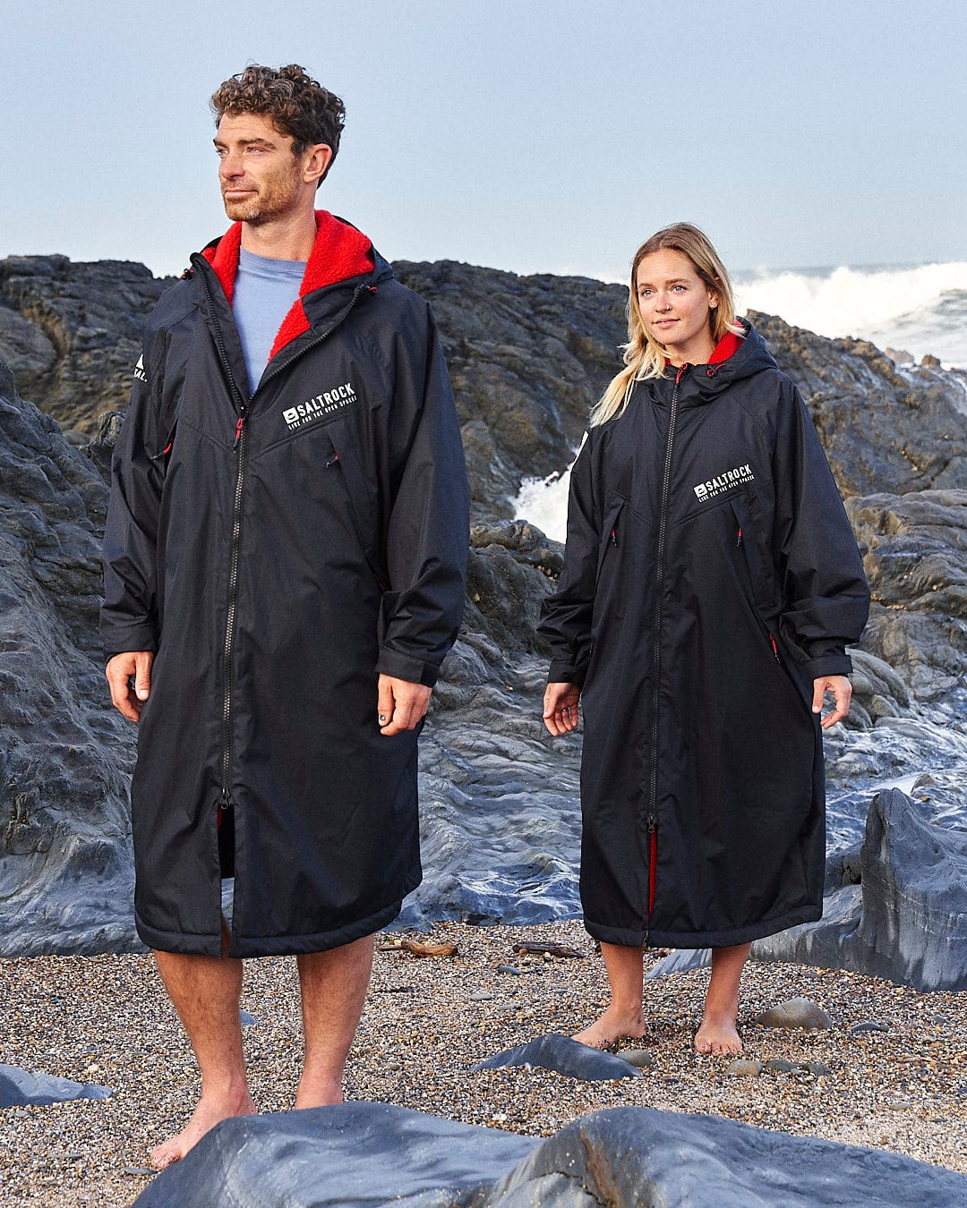 Two people stand barefoot on a rocky beach, each donning a long black waterproof Recycled Changing Robe - Black/Red made from recycled materials. Waves crash in the background as the Saltrock logo is proudly displayed on the upper chest of their jackets.