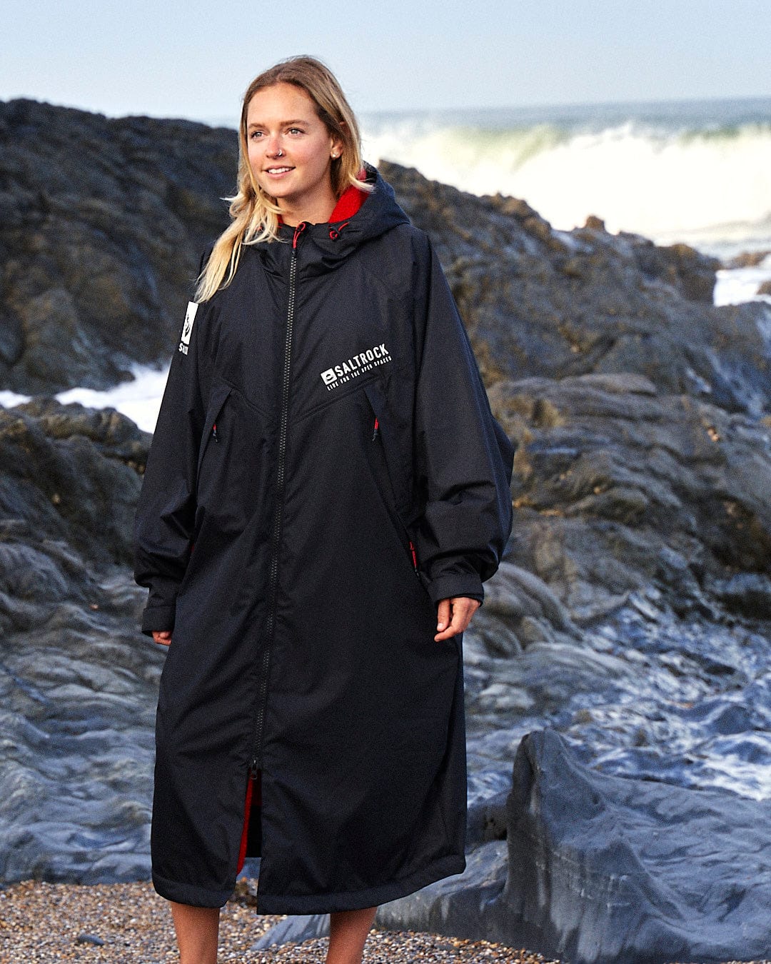 A person wearing a long, black Recycled Changing Robe - Black/Red with the Saltrock logo stands on a rocky beach with waves crashing in the background.