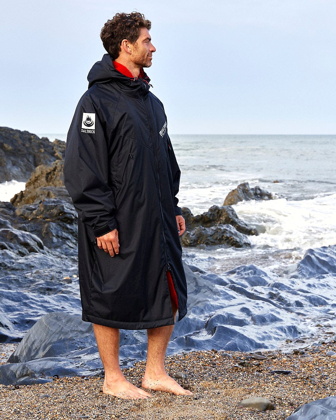 A person with short curly hair stands barefoot on a rocky beach, wearing a Saltrock Recycled Changing Robe - Black/Red made from recycled materials with patches on the sleeves. The ocean waves can be seen in the background, and a subtle Saltrock logo is visible on the hood.