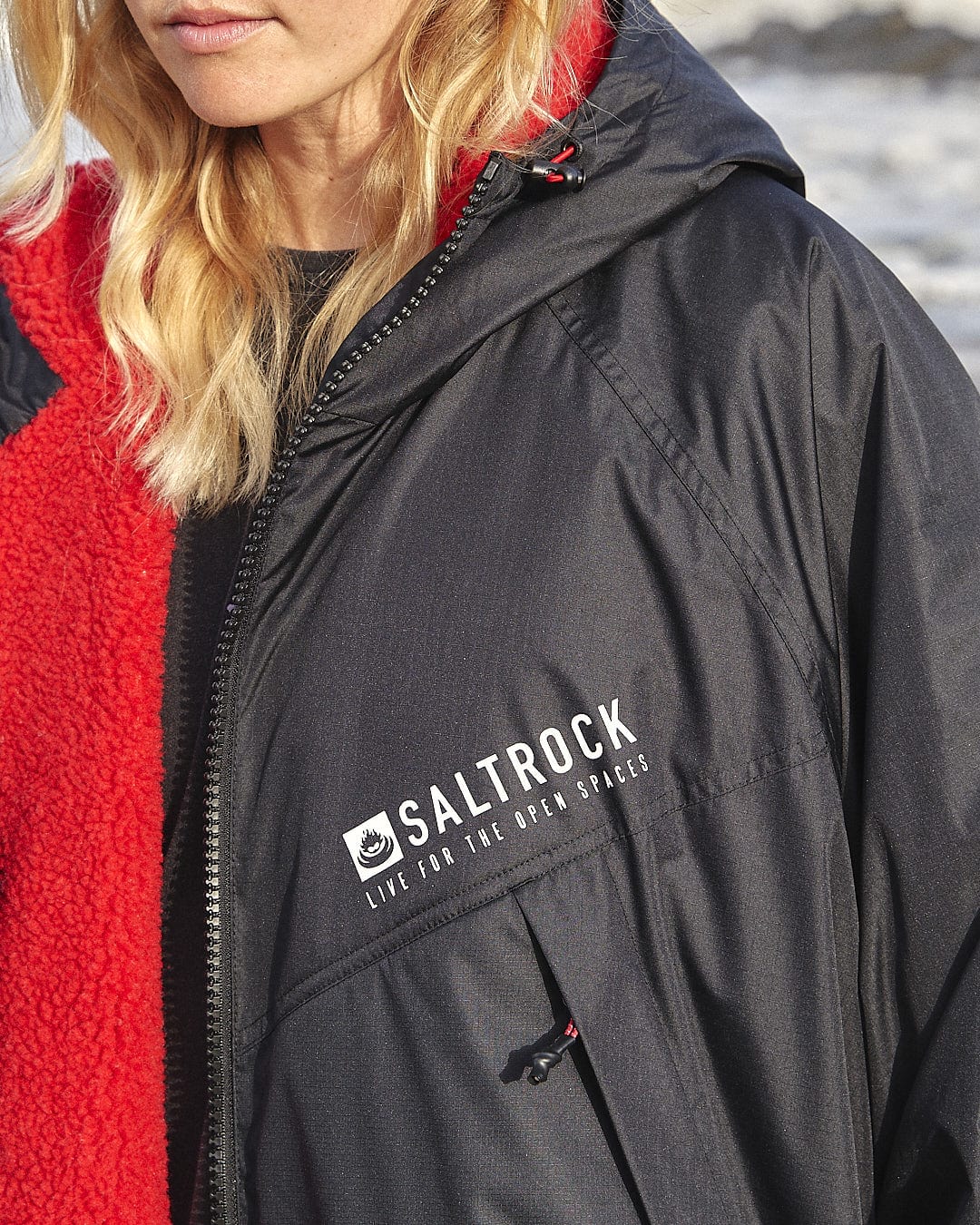 Close-up of a person wearing a black Saltrock Recycled Changing Robe - Black/Red featuring the iconic "Live for the Open Spaces" slogan and a red, textured garment underneath. The person, with blonde hair, showcases the Saltrock logo prominently.