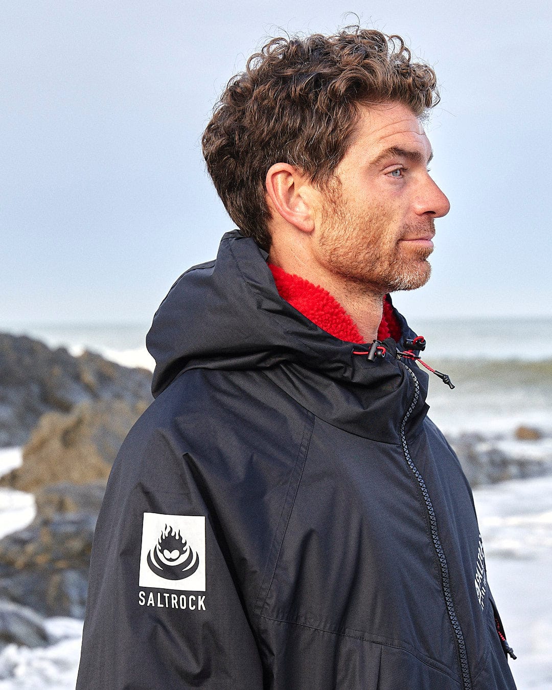 A man with curly hair wearing a Saltrock Recycled Four Seasons Changing Robe in Black/Red gazes into the distance by a rocky beach.