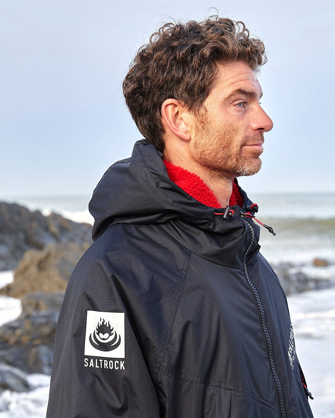 A man in a dark jacket with a "Saltrock" logo stands on a rocky beach, showcasing the Saltrock Recycled Changing Robe - Black/Red crafted from recycled materials.