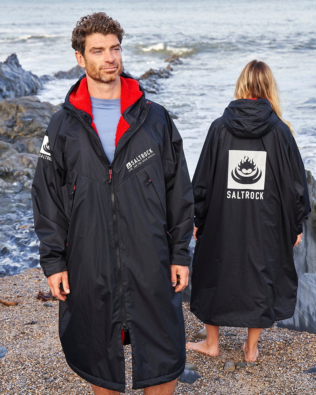Two people stand on a rocky beach wearing black Saltrock Recycled Changing Robes - Black/Red, made from recycled materials. One faces forward, and the other turns away, both near the ocean. The Saltrock logo is visible on their robes as they embrace the coastal breeze.
