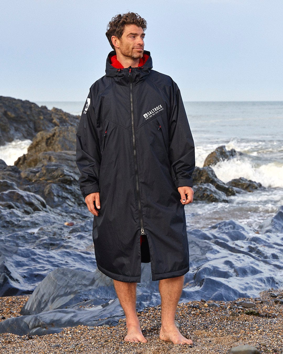 A man stands barefoot on a rocky beach, wearing a Saltrock Recycled Changing Robe - Black/Red with a hood made from recycled materials. Waves are crashing in the background.