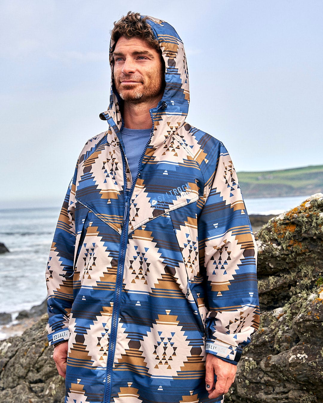A man wearing a Saltrock Four Seasons Changing Robe - Aztec print raincoat standing on a rocky beach.