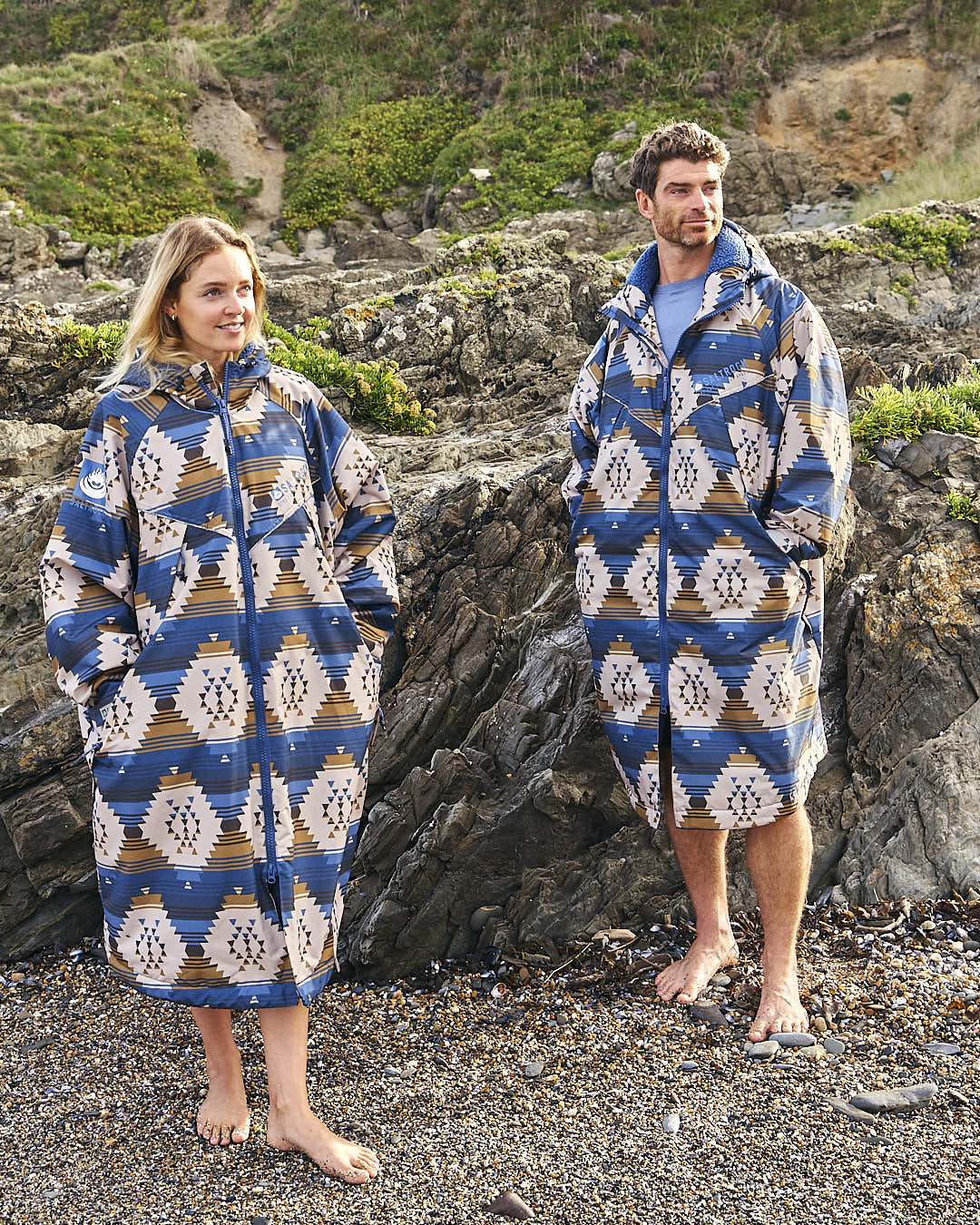 A man and woman standing on a rocky beach wearing the Saltrock Four Seasons Changing Robe - Aztec.