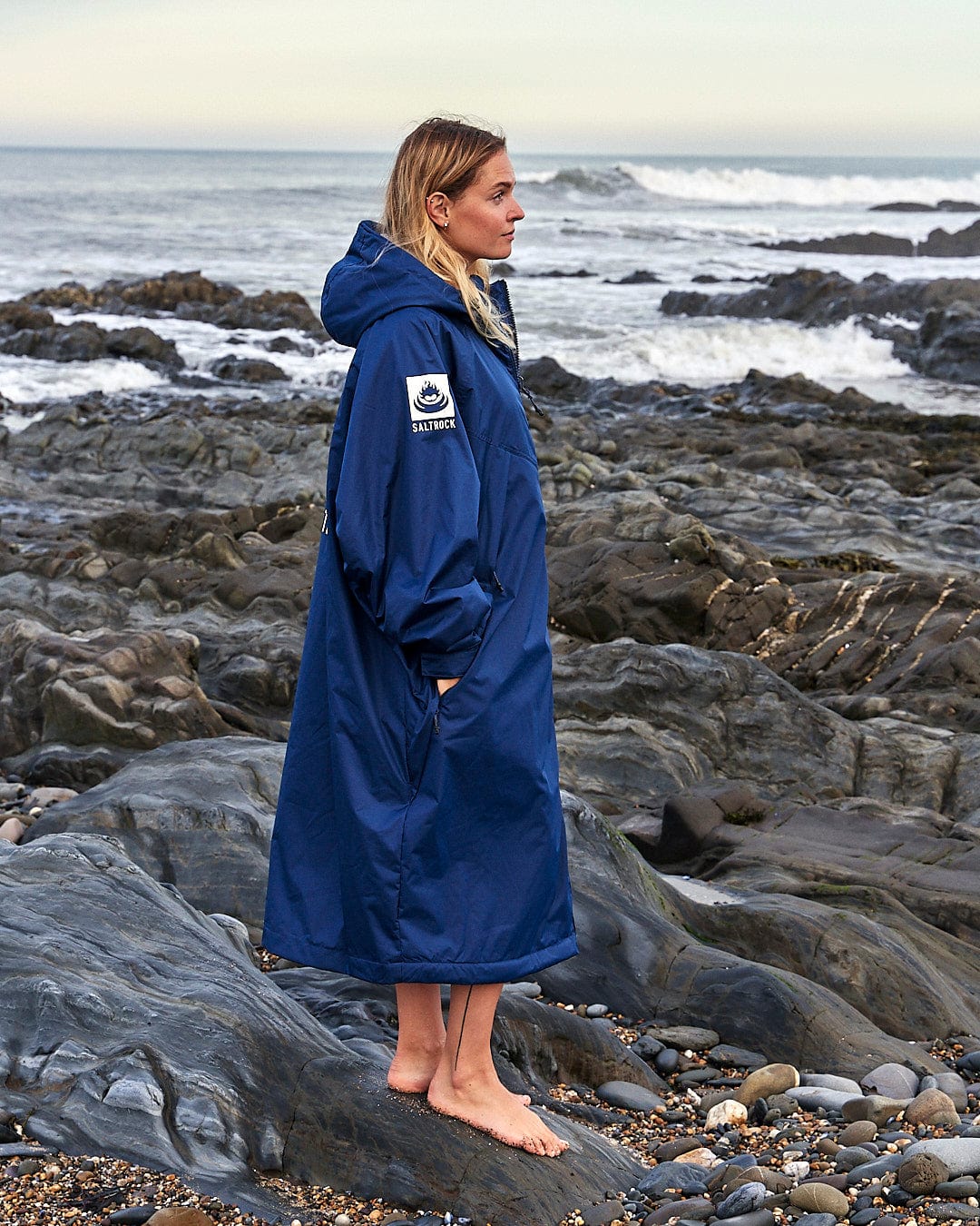 A person in a Saltrock Changing Robe - Blue stands barefoot on the rocky shore with ocean waves in the background.