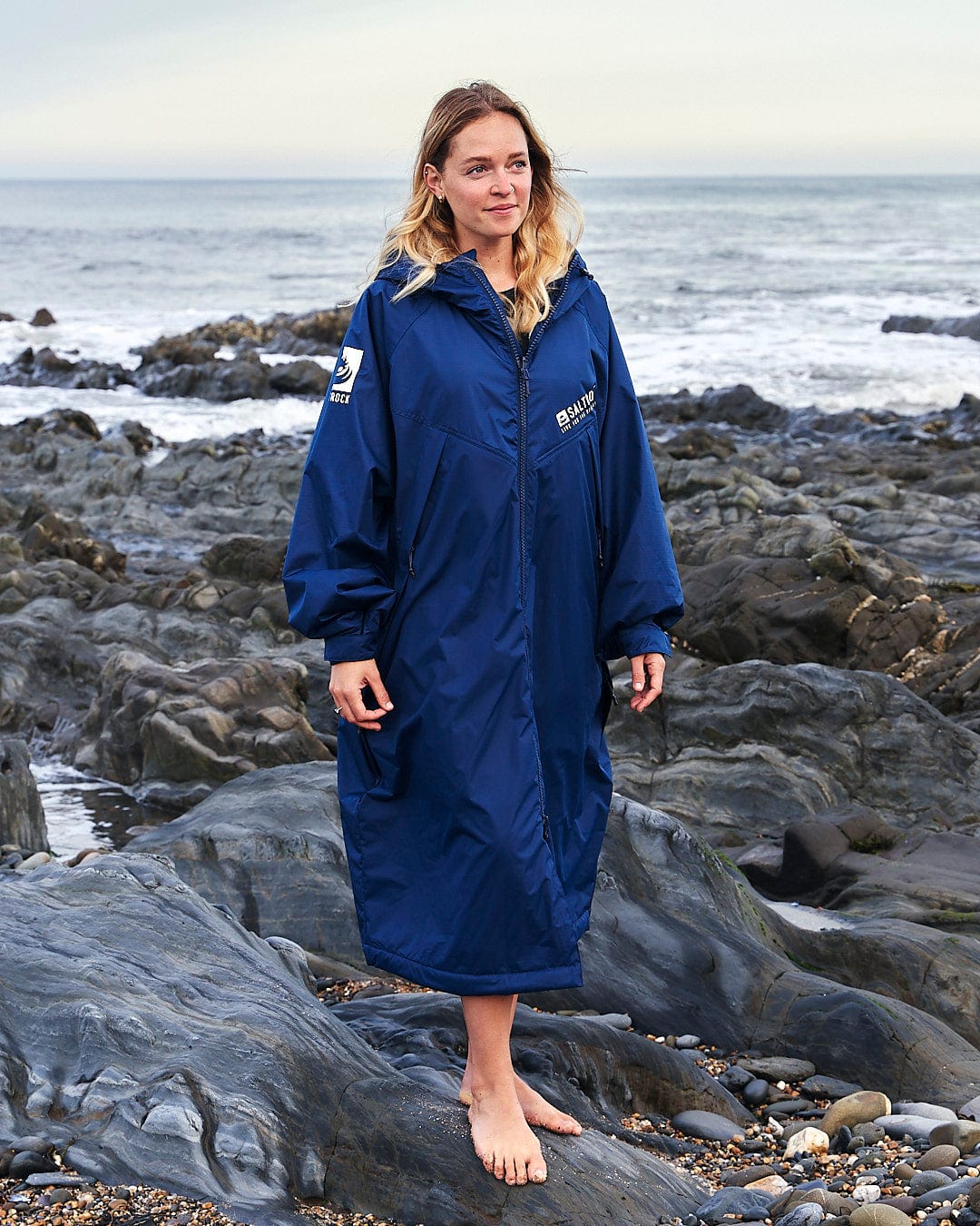 A woman, clad in a Changing Robe - Blue with the Saltrock logo, stands on rocks near the water.