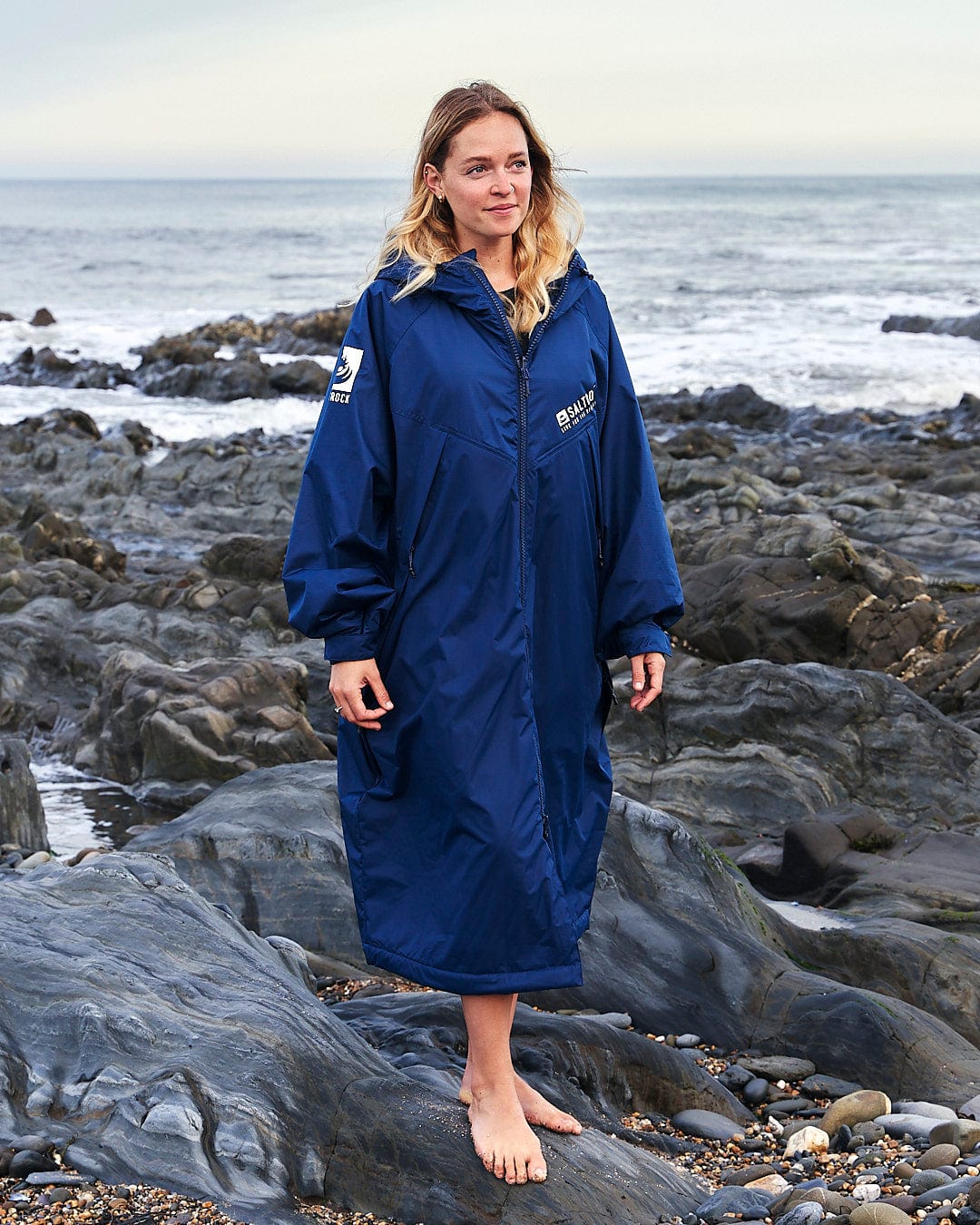 A woman stands barefoot on a rocky beach, wearing a large blue Saltrock Recycled Four Seasons Changing Robe, looking off into the distance with a thoughtful expression.