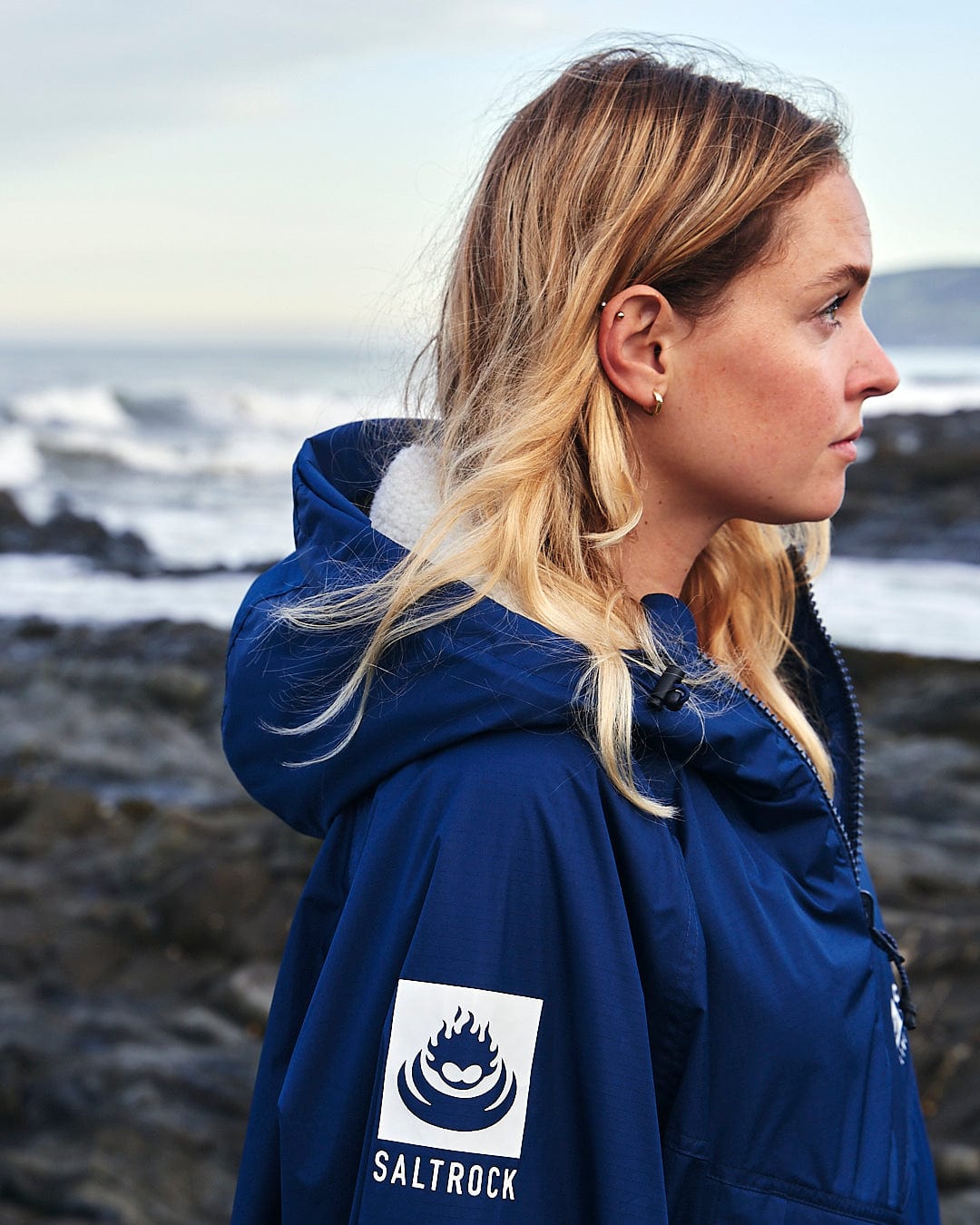 A woman with shoulder-length blonde hair wearing a blue Saltrock Changing Robe - Blue, adorned with the Saltrock logo, stands on a rocky beach with the sea in the background.