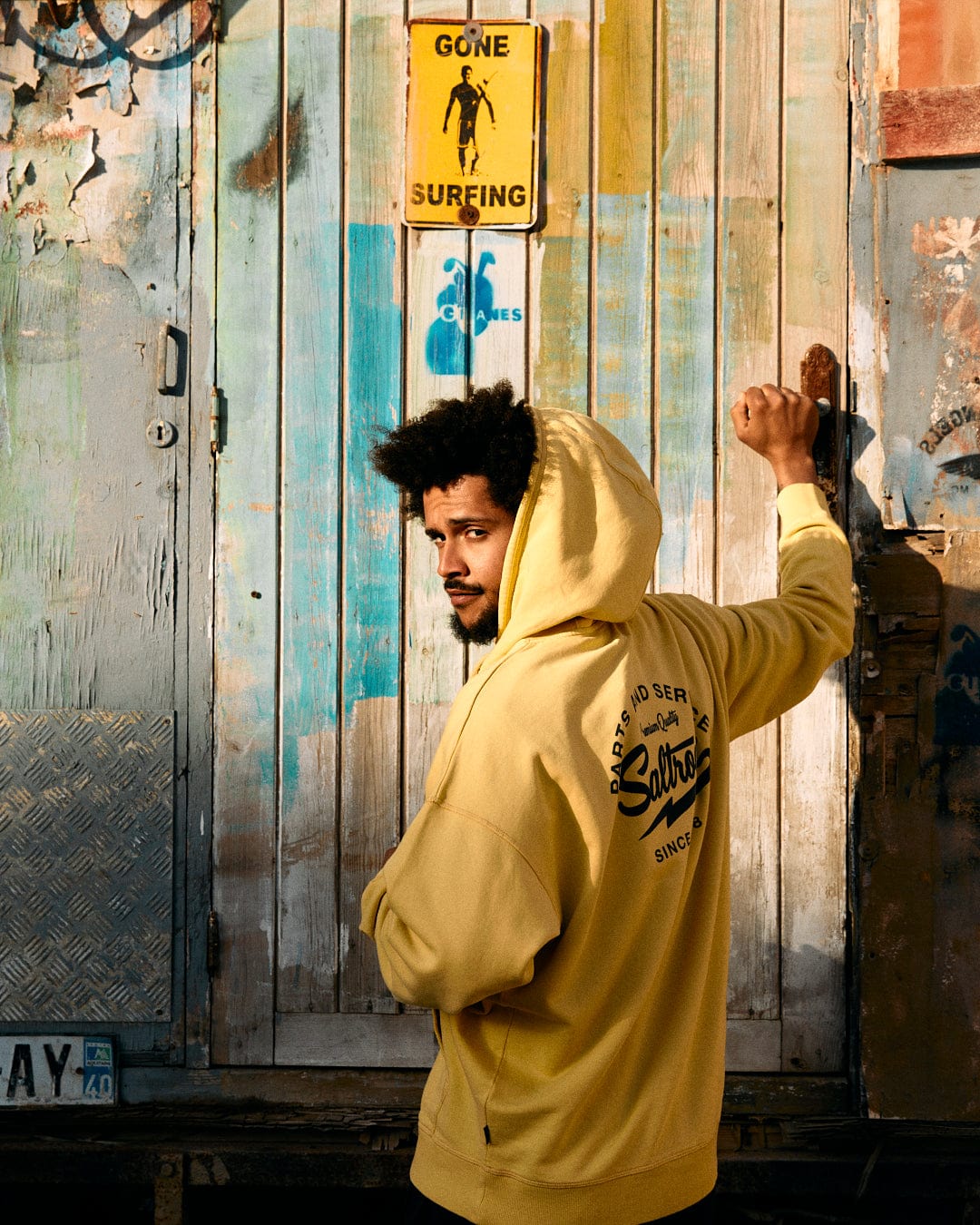 A man in a yellow Saltrock Gas Station - Recycled Mens Pop Hoodie stands in front of a rustic blue door, looking over his shoulder, under a yellow sign depicting a figure.