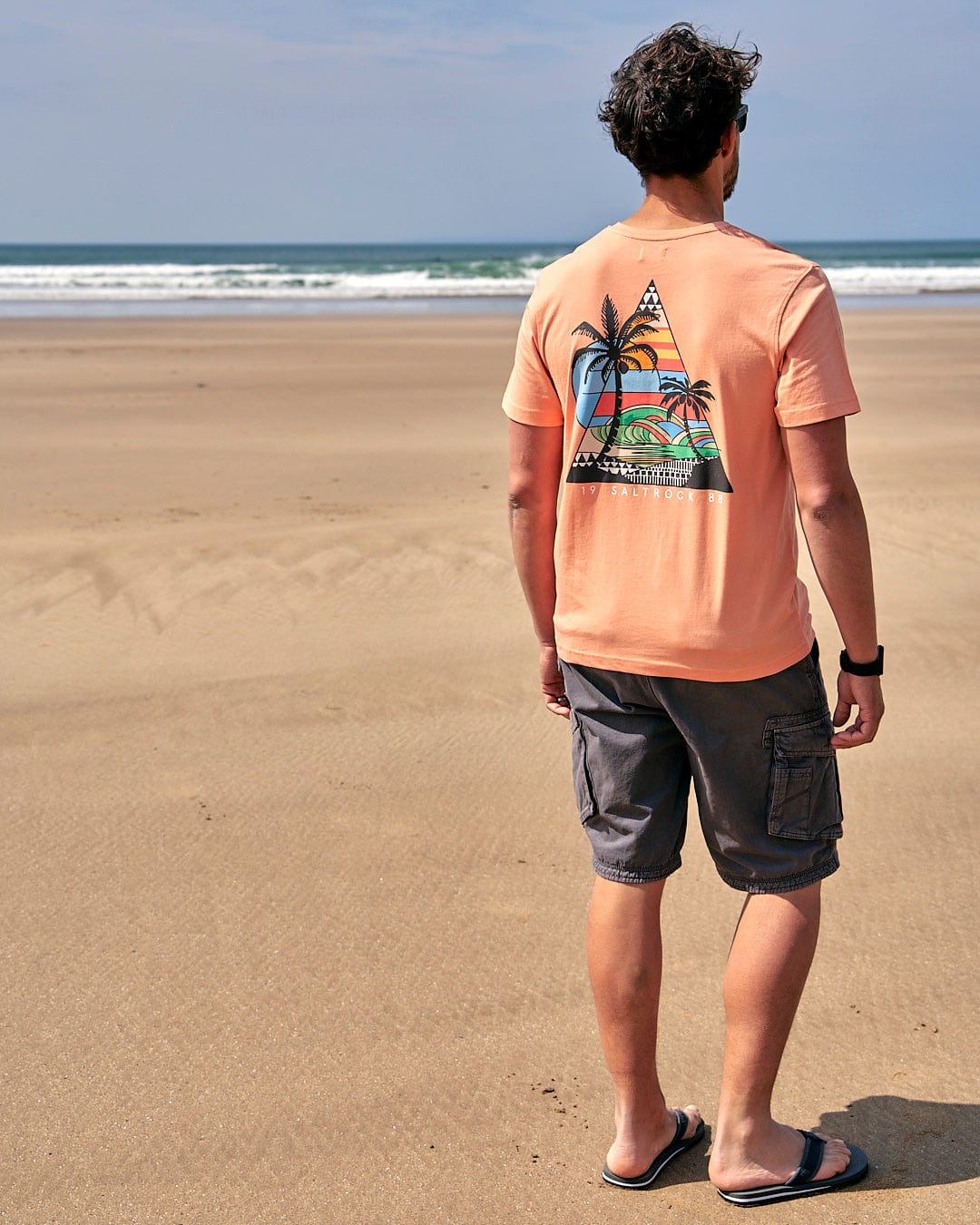 A man is standing on the beach wearing a Saltrock Geo Beach - Mens Short Sleeve T-Shirt - Coral.