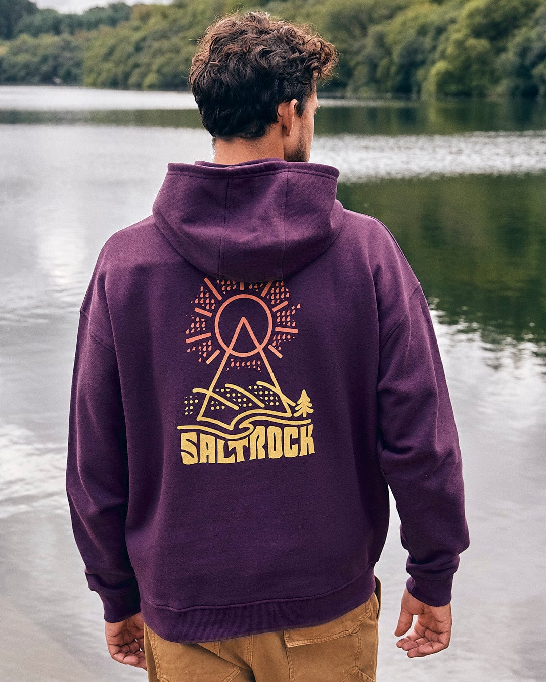 A man is standing by a lake wearing a Saltrock Geo Peak - Mens Oversized Snood Pop Hoodie - Dark Purple.