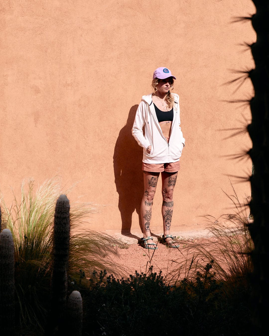 Woman in a white jacket and cap, featuring Saltrock branding, standing beside cacti against a peach-colored wall, sunglasses perched atop her head wearing the Ginny - Womens Zip Hoodie - Cream by Saltrock.