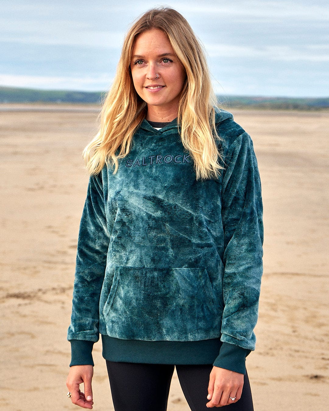 A woman wearing a Saltrock Gracie - Womens Tie Dye Velour Pop Hoodie - Dark Green standing on the beach.