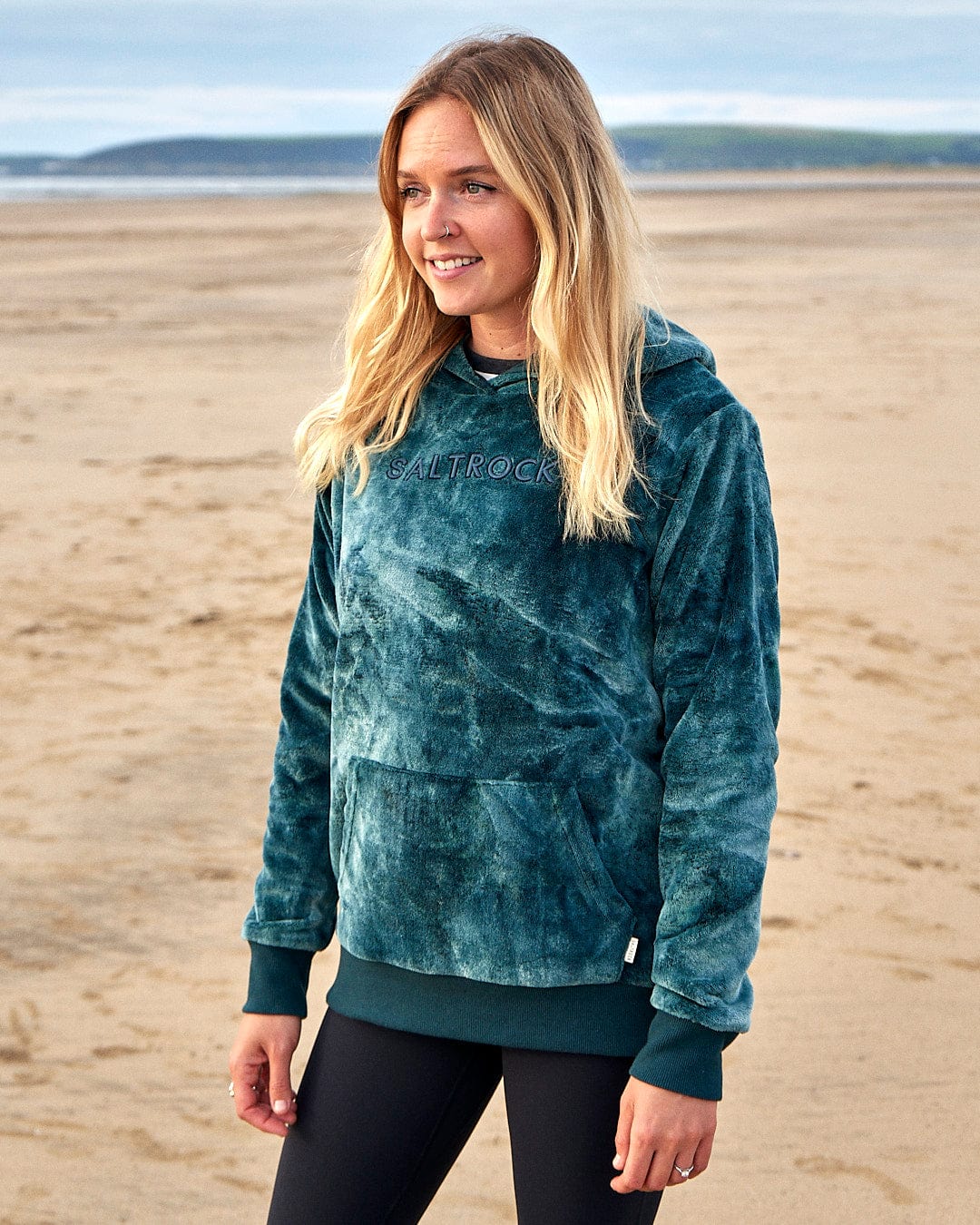 A woman wearing the Saltrock Gracie - Womens Tie Dye Velour Pop Hoodie - Dark Green on the beach.