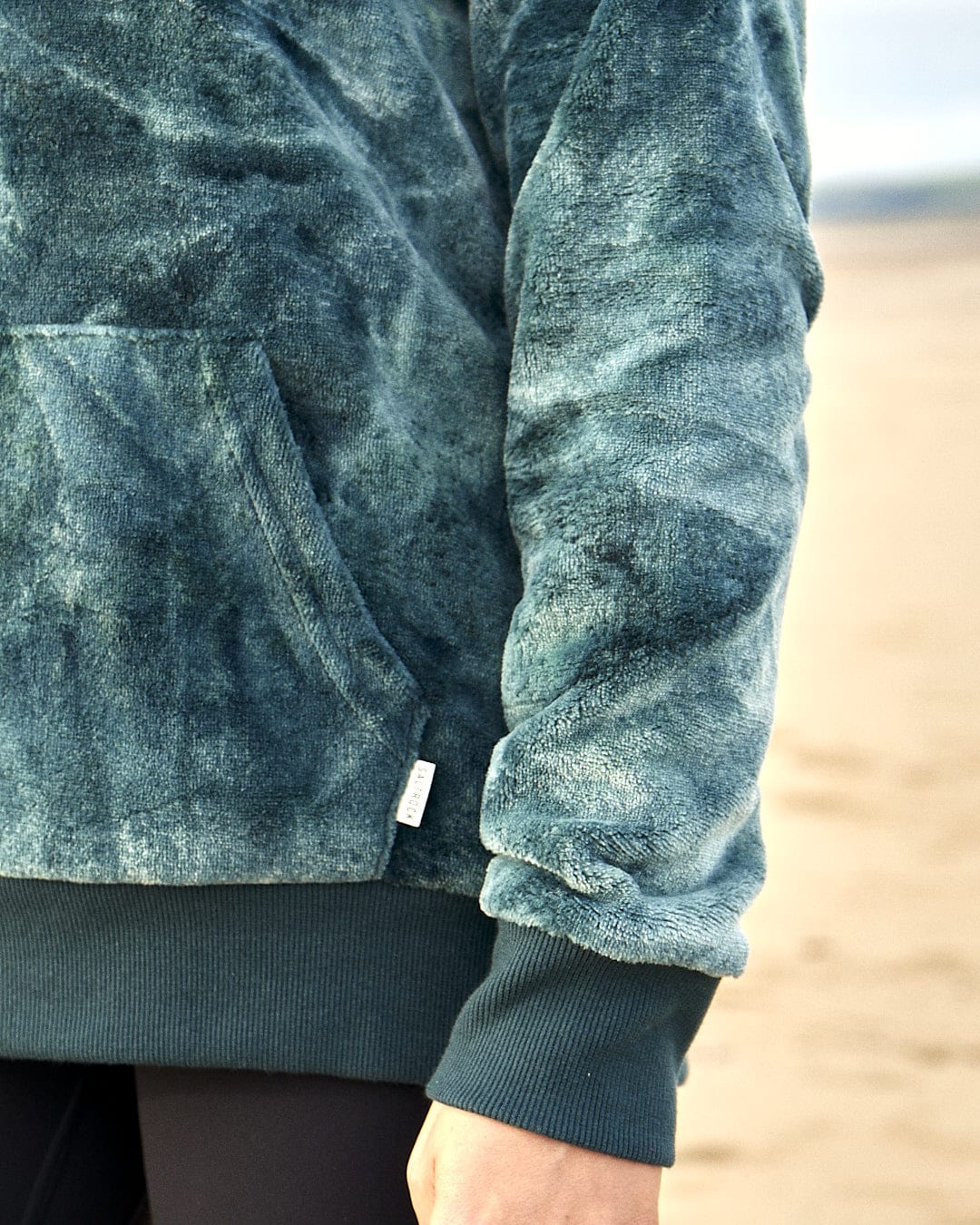 A woman wearing a Saltrock Gracie - Womens Tie Dye Velour Pop Hoodie - Dark Green on the beach.
