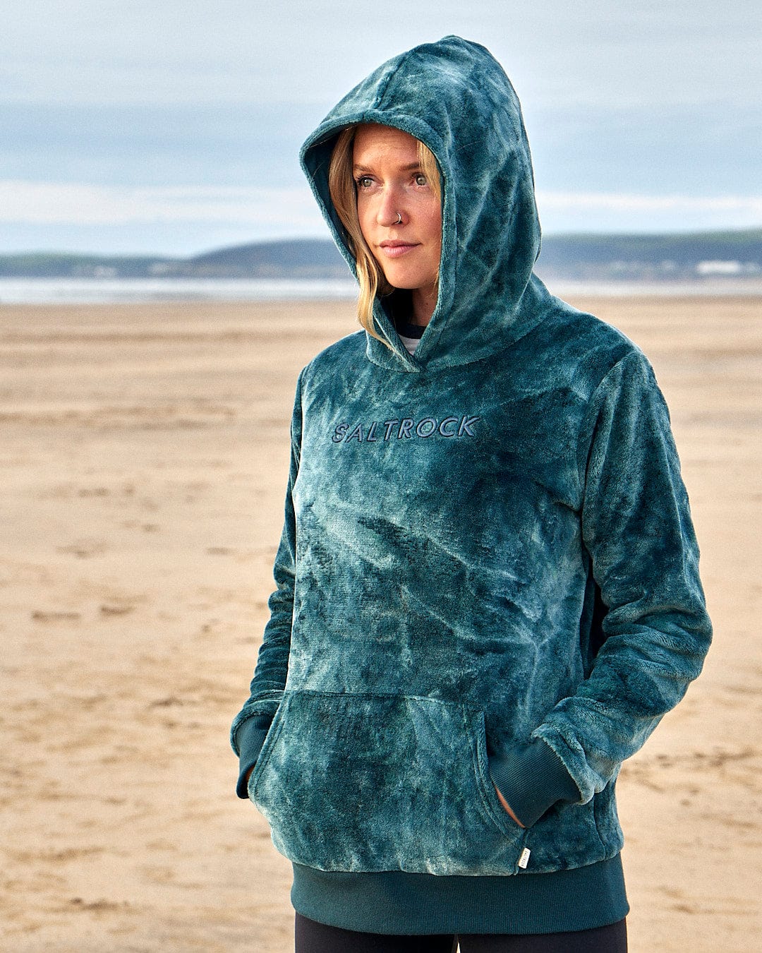 A woman wearing a Saltrock Gracie - Womens Tie Dye Velour Pop Hoodie - Dark Green standing on the beach.