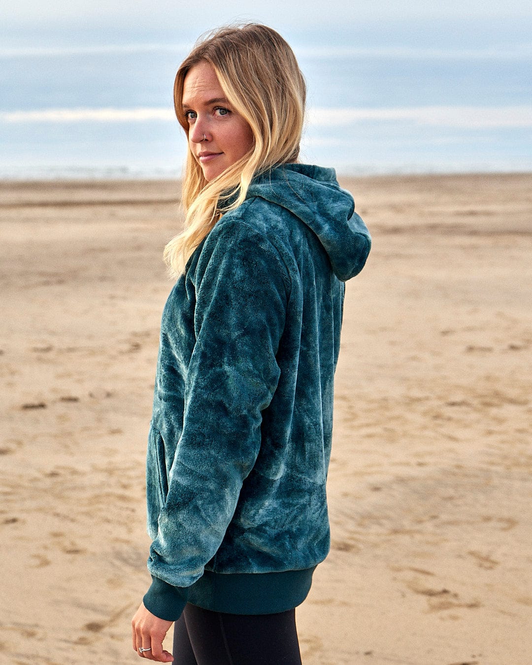 A woman wearing a Saltrock Gracie - Womens Tie Dye Velour Pop Hoodie - Dark Green on the beach.