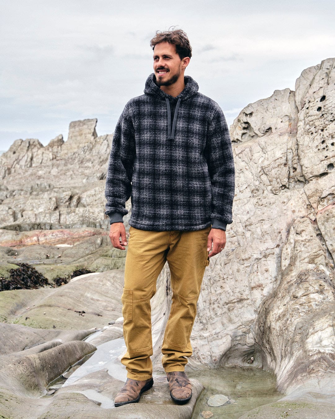 A man standing on rocks with a Saltrock Halycon - Mens Fleece Hoodie - Dark Grey made of fleece material.