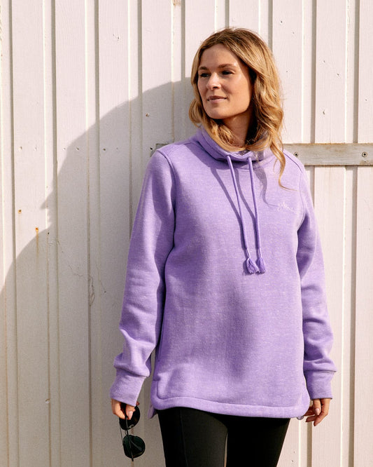 Person standing in front of a white wooden wall wearing the Saltrock Harper - Womens Longline Pop Sweat in Lilac and holding sunglasses.