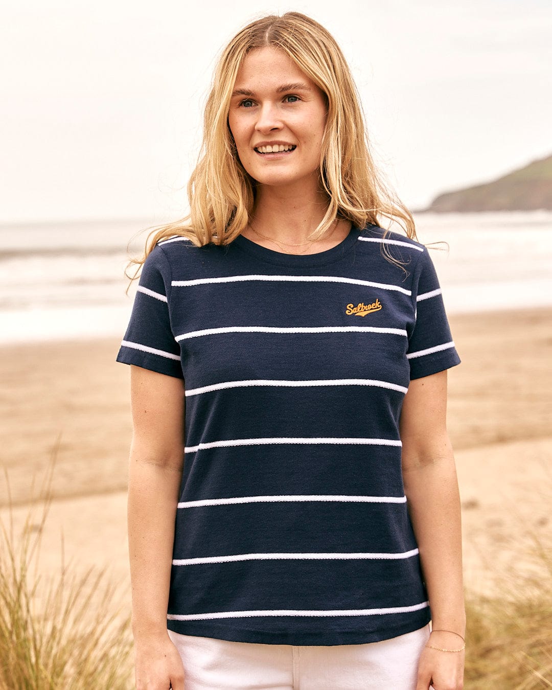 A woman wearing a Saltrock Hartland - Womens Striped Short Sleeve T-Shirt - Blue standing on the beach.