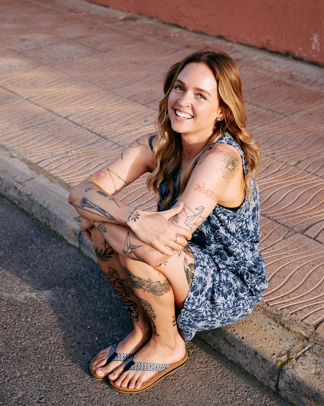 A young woman with tattoos sits smiling on a sidewalk, wearing a Hibiscus Bauhaus - Womens Midi Dress in Blue by Saltrock and flip-flops, during golden hour.