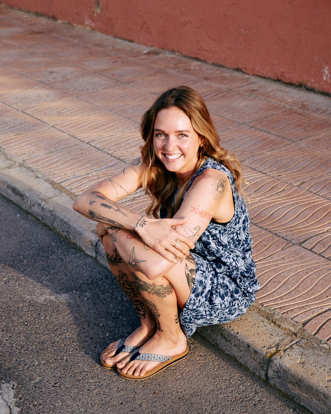 A woman with tattoos smiles while sitting on a sidewalk, wearing a blue Saltrock branded Hibiscus Bauhaus - Womens Midi Dress and flip-flops.