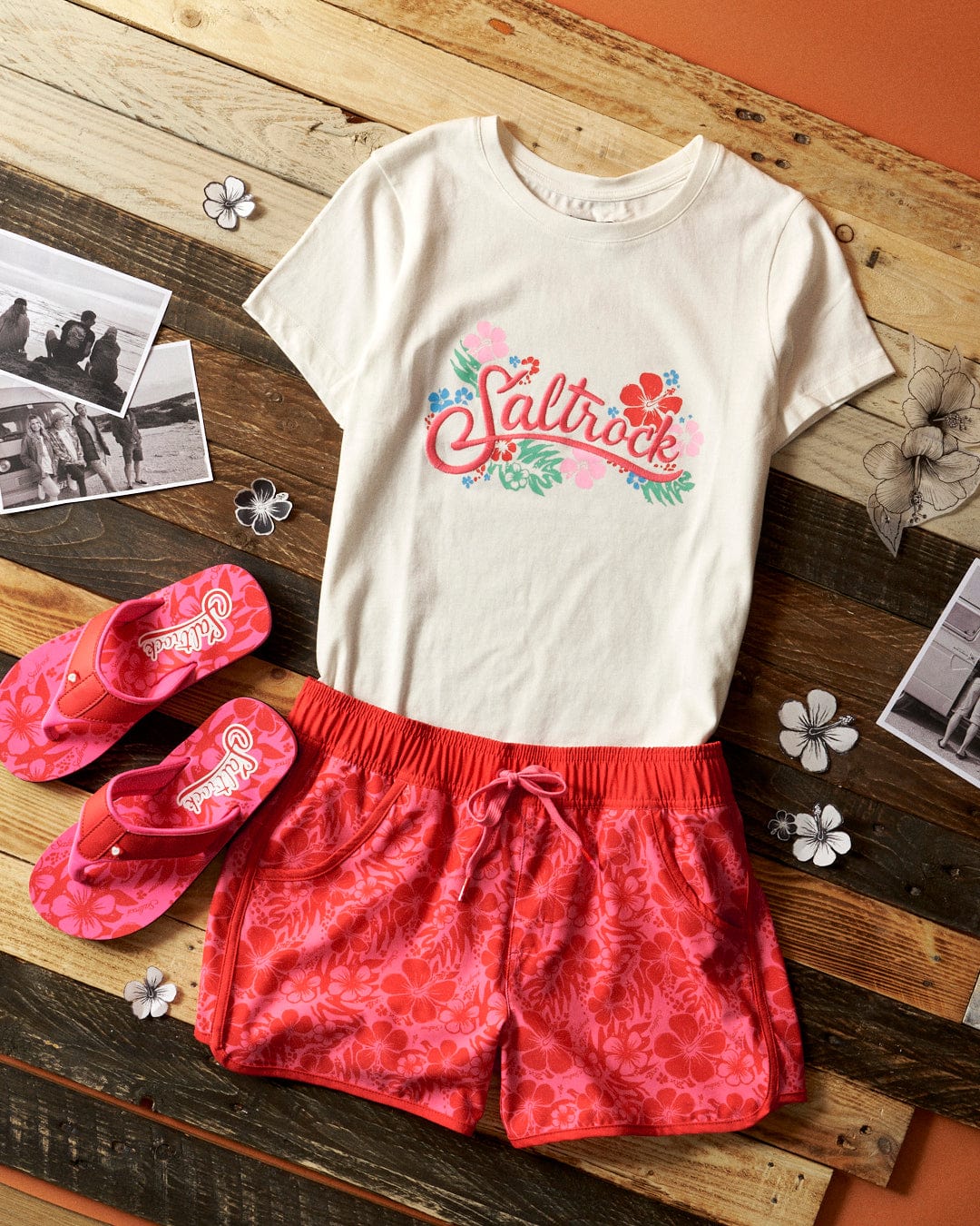 A t-shirt, Hibiscus - Womens Boardshorts - Red/Pink, and flip flops on a wooden table by Saltrock.