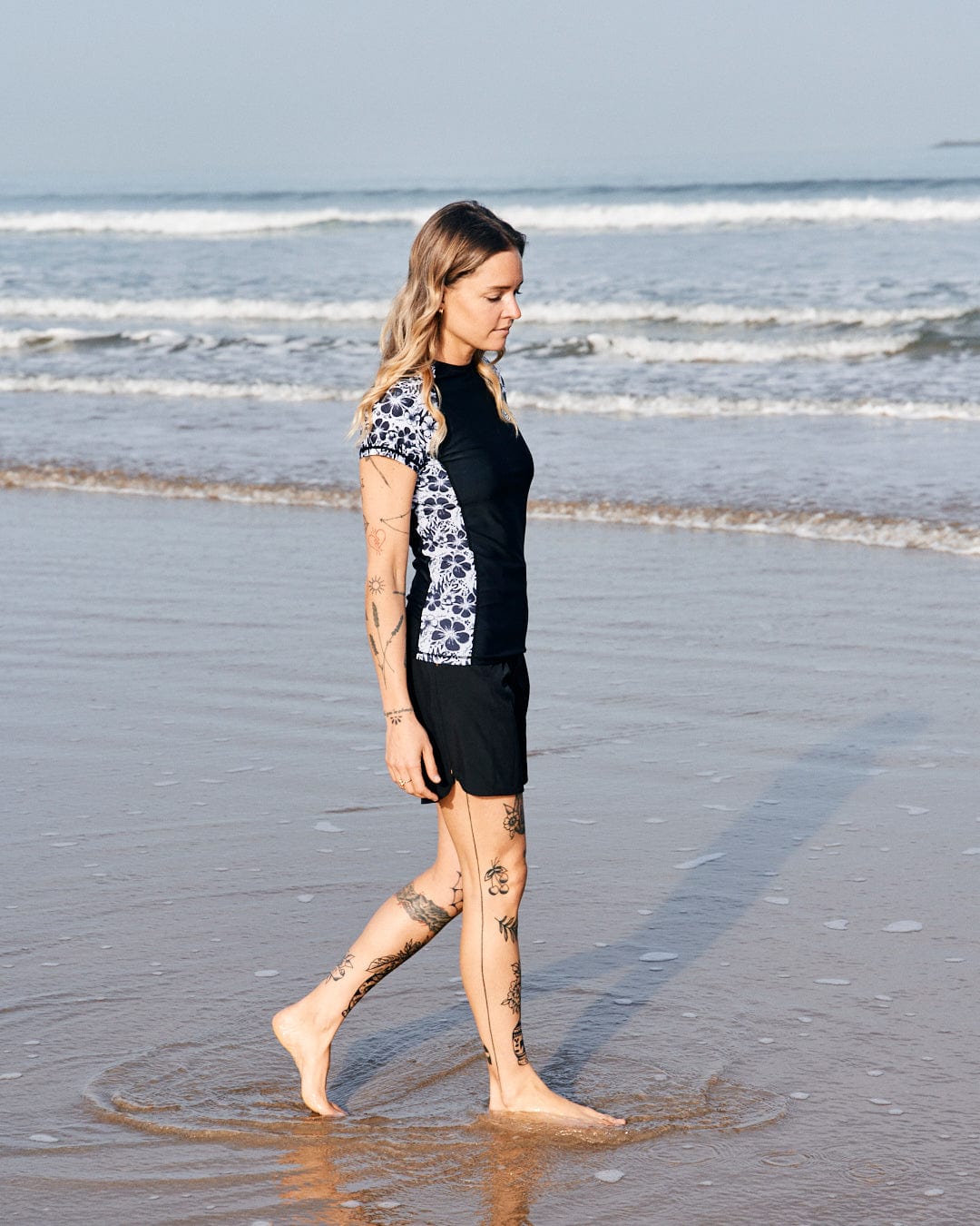 A woman with tattoos walks barefoot on a sandy beach, wearing a black and white Saltrock Hibiscus - Recycled Womens Short Sleeve Rashvest - Black shirt and shorts, with ocean waves in the background.