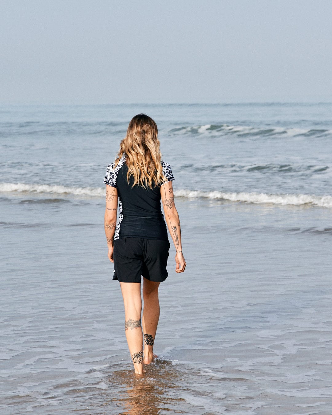 A woman with tattoos walks into the sea, wearing a black outfit with Saltrock Hibiscus - Recycled Womens Short Sleeve Rashvest - Black pattern, viewed from behind.