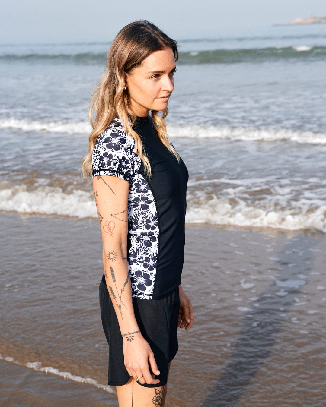 A woman with tattoos stands on a beach, wearing a blue and white rash guard featuring the Saltrock Hibiscus - Recycled Womens Short Sleeve Rashvest - Black, looking sideways with the ocean in the background.