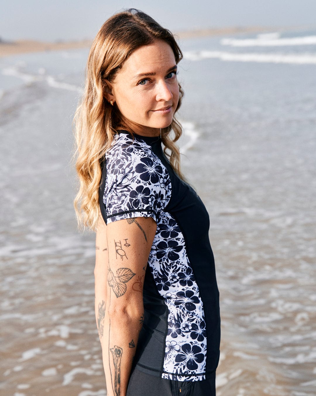 Woman with tattoos wearing a Hibiscus - Recycled Womens Short Sleeve Rashvest - Black by Saltrock, featuring UPF 50 protection, standing on a beach with waves in the background.