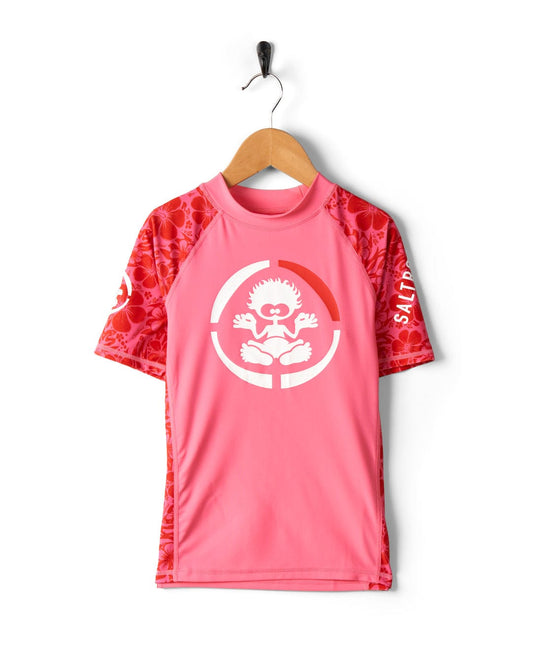 Hibiscus - Recycled Kids Short Sleeve Rashvest - Pink athletic t-shirt with red patterned sleeves hanging on a hook against a white background, featuring a circular logo with a heart and hands design and UPF 50 protection. (Saltrock)