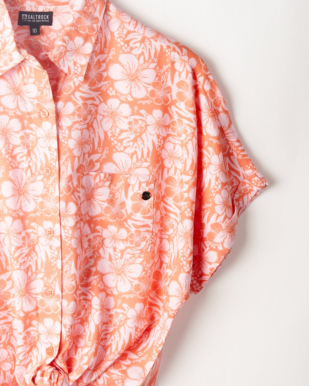 A close-up of a coral-colored hibiscus floral print shirt with a black button, displaying the label "Saltrock" at the collar.