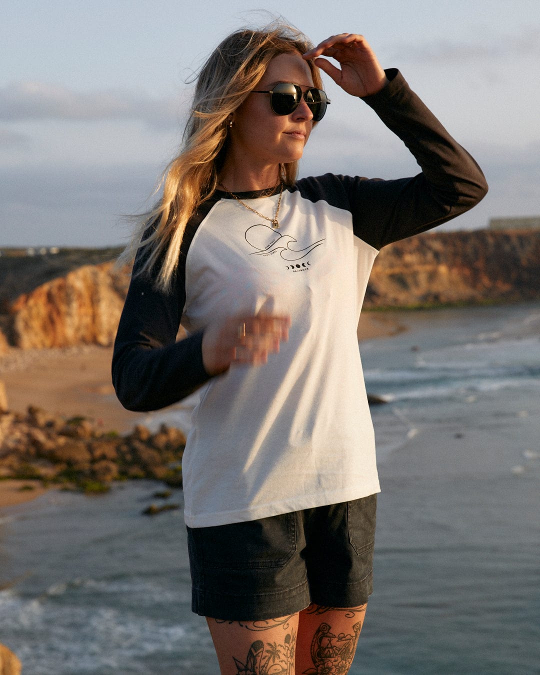 A woman with graphic tattoos standing on a cliff overlooking the ocean wearing the Saltrock High Tides - Womens Raglan Long Sleeve T-Shirt in White.
