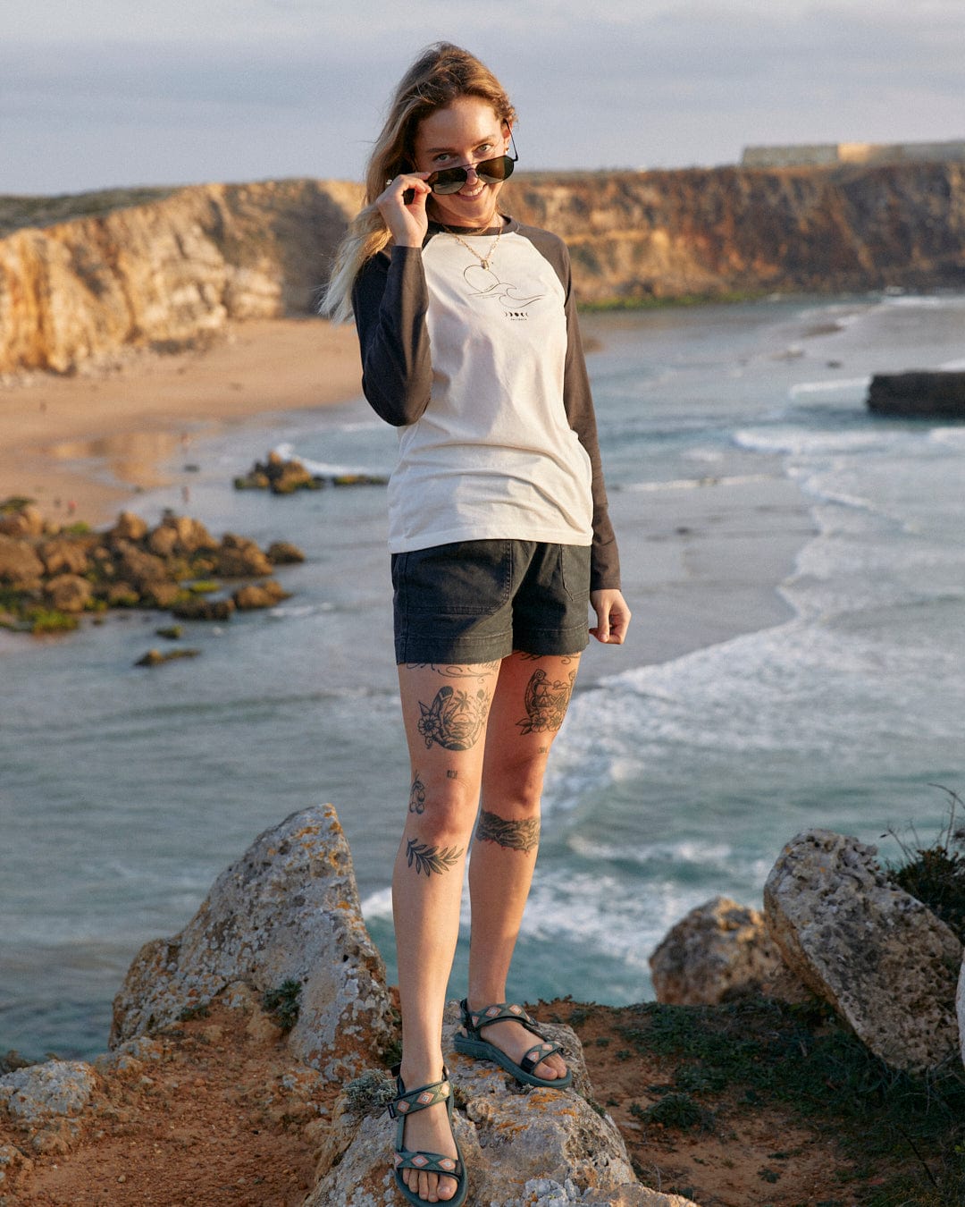 A woman with tattoos standing on a rock by the ocean wearing Saltrock's High Tides - Womens Raglan Long Sleeve T-Shirt in White.