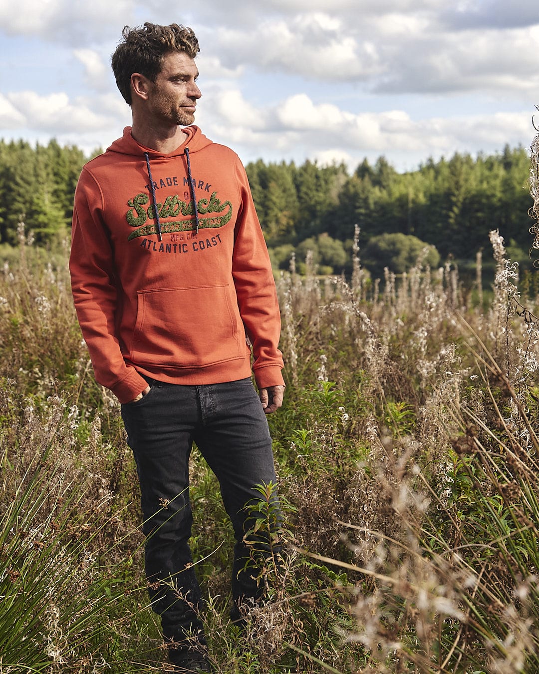 A man wearing an orange Saltrock Home Run Chenille - Mens Pop Hoodie - Red stands in a field.
