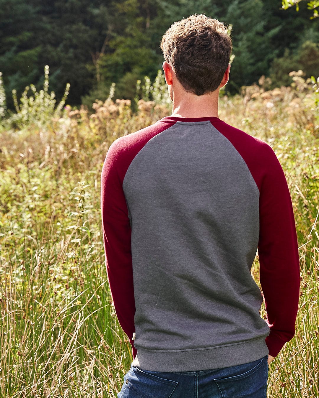 A sporty man wearing a Saltrock Home Run - Mens Long Sleeve Sweat - Grey/Red with contrasting red sleeves stands in a field.