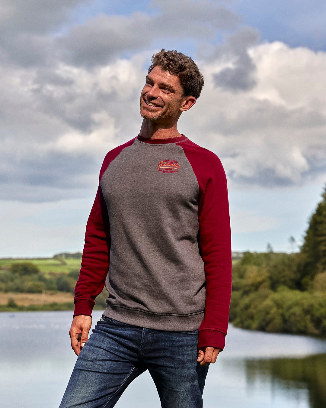 A man in a Saltrock Home Run - Mens Long Sleeve Sweat - Grey/Red with contrasting red sleeves standing next to a lake.