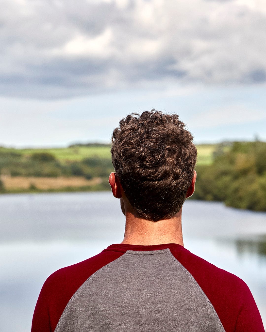 A man wearing a Saltrock Home Run - Mens Long Sleeve Sweat - Grey/Red with contrasting red sleeves gazes out over a lake.
